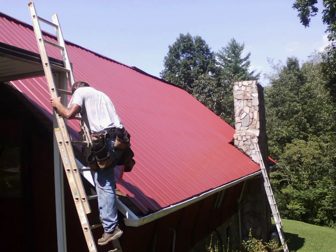 Roofer on a ladder beside a red metal roof and stone chimney — Soffit & Fascia Repair by Roofing Brothers in Pascoag
